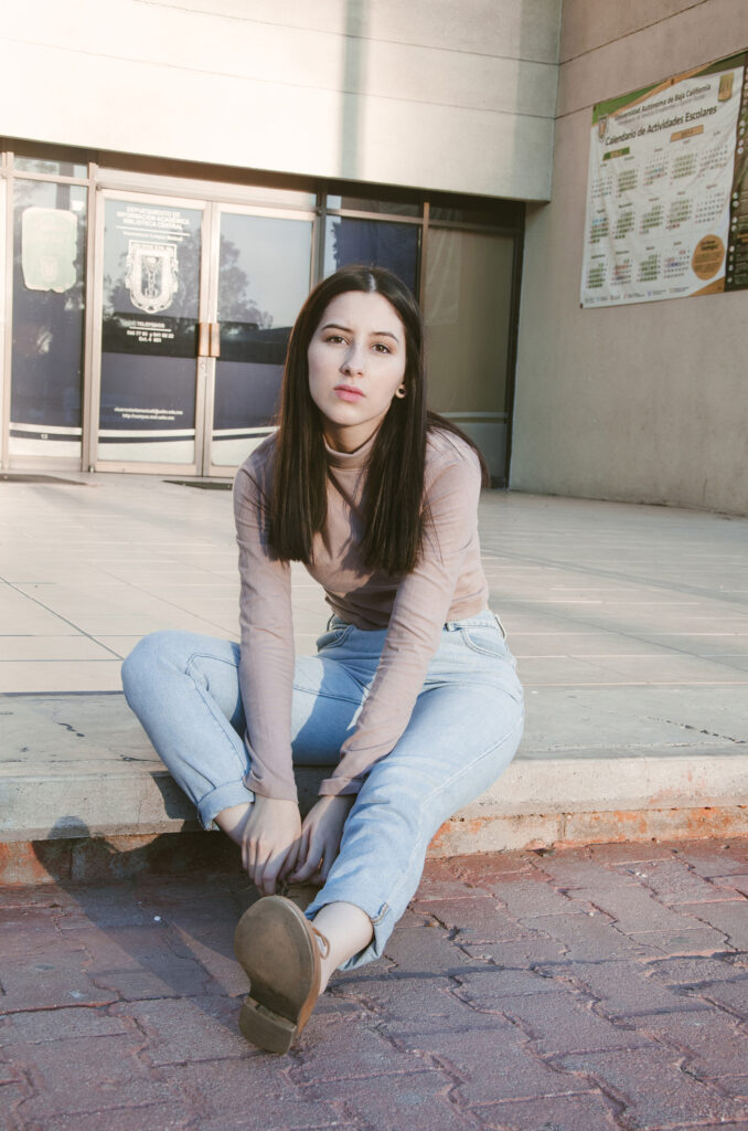 Gabriela Inzunza sits on a brick pathway outside a building with a calendar on the wall wearing a beige top and blue jeans.